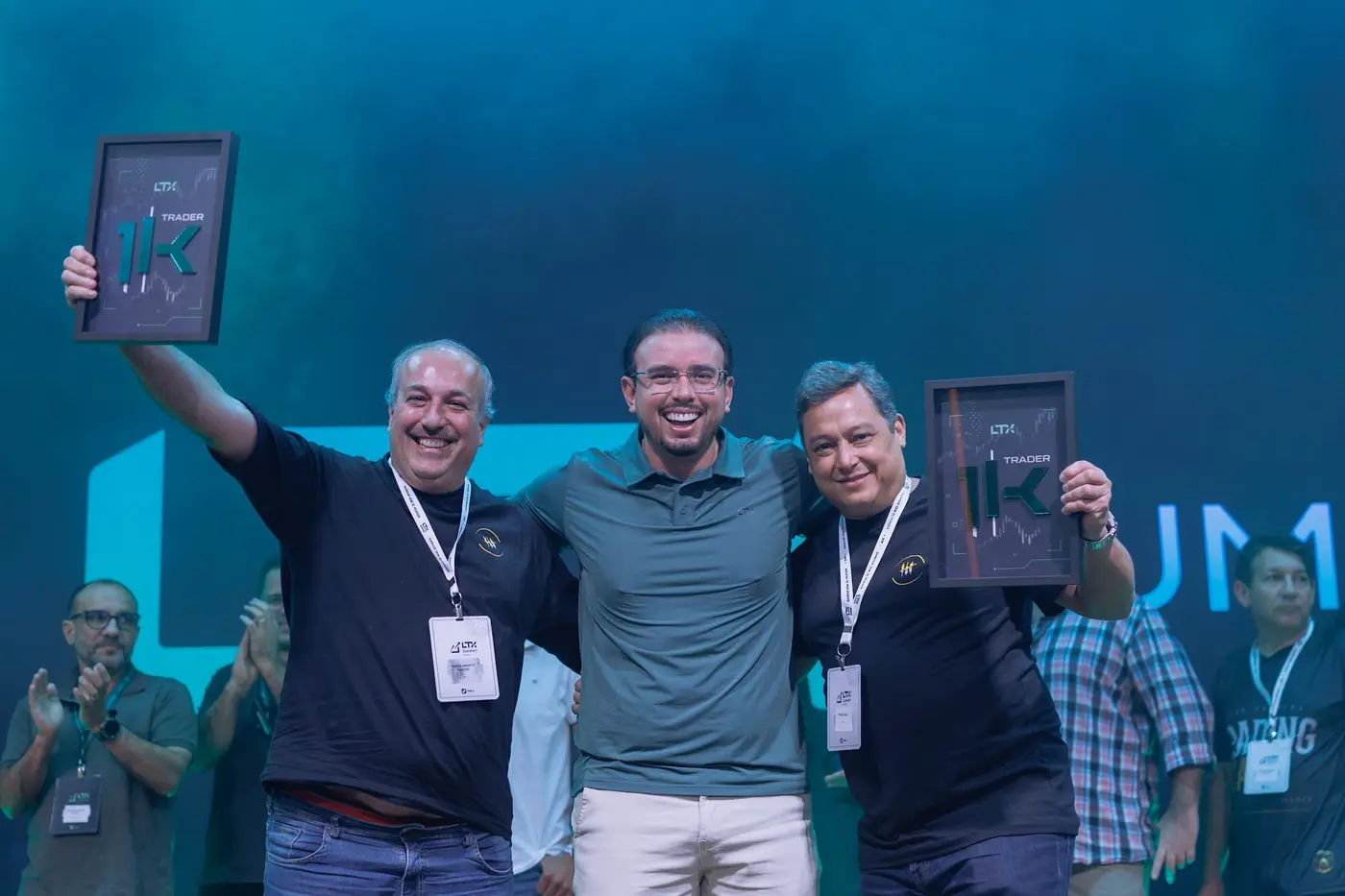 Três traders comemorando no palco com a placa Trader 1K