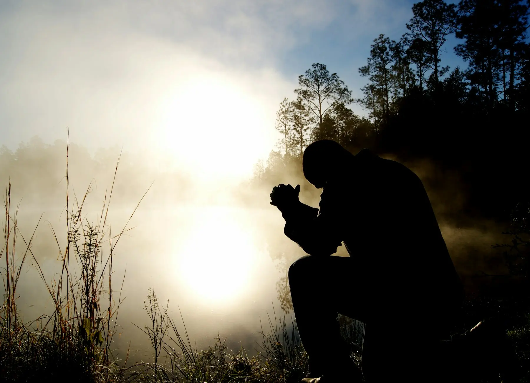 Silhueta de um homem ajoelhado orando ao amanhecer, com névoa e luz do sol