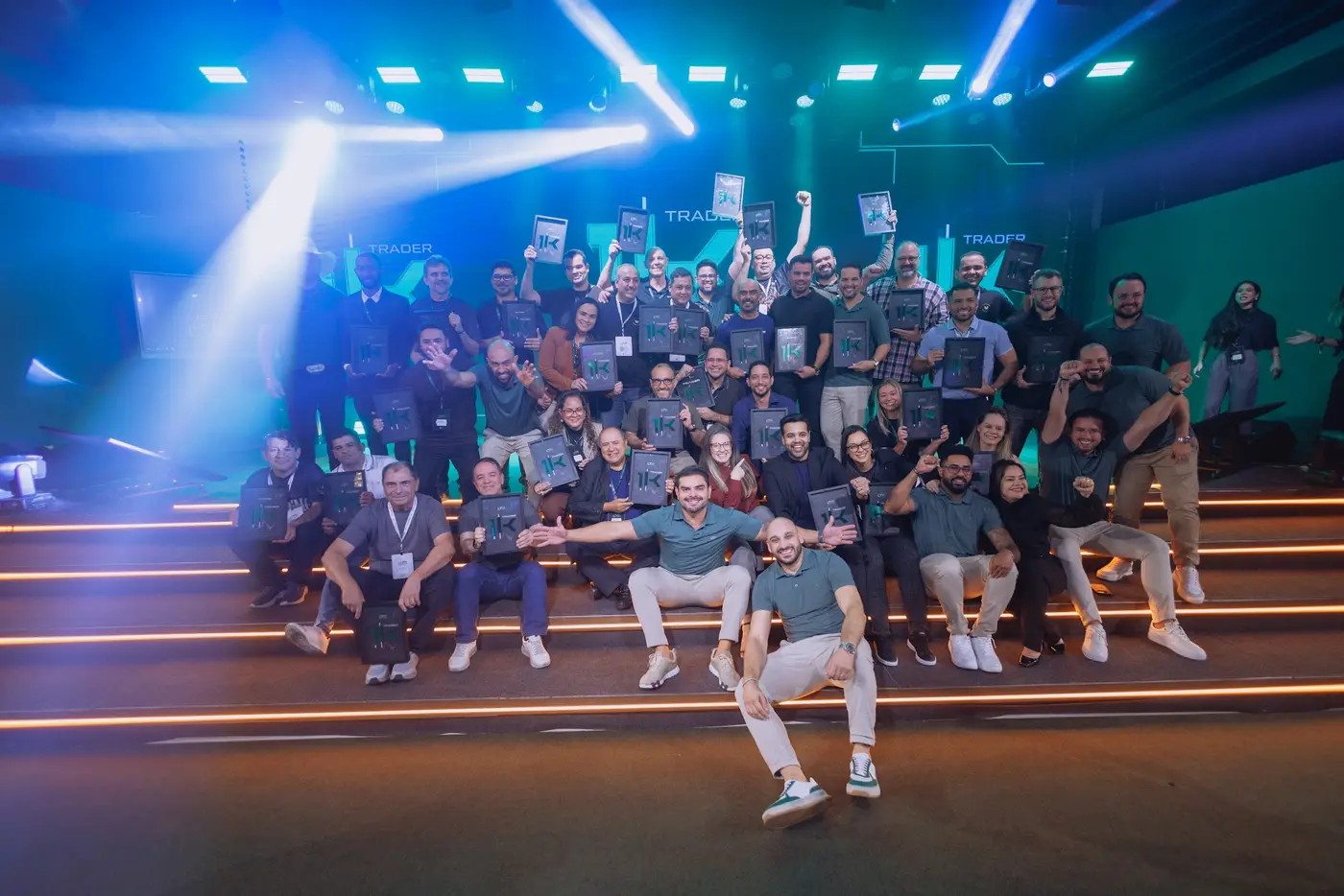 Turma Trader 1K reunida nos degraus do palco com feixes de luz