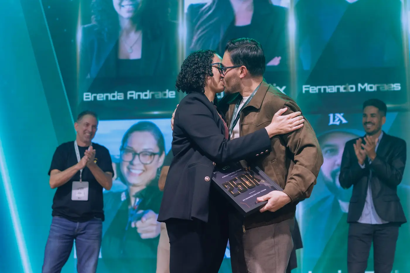 Casal se beijando no palco segurando a placa de conquista