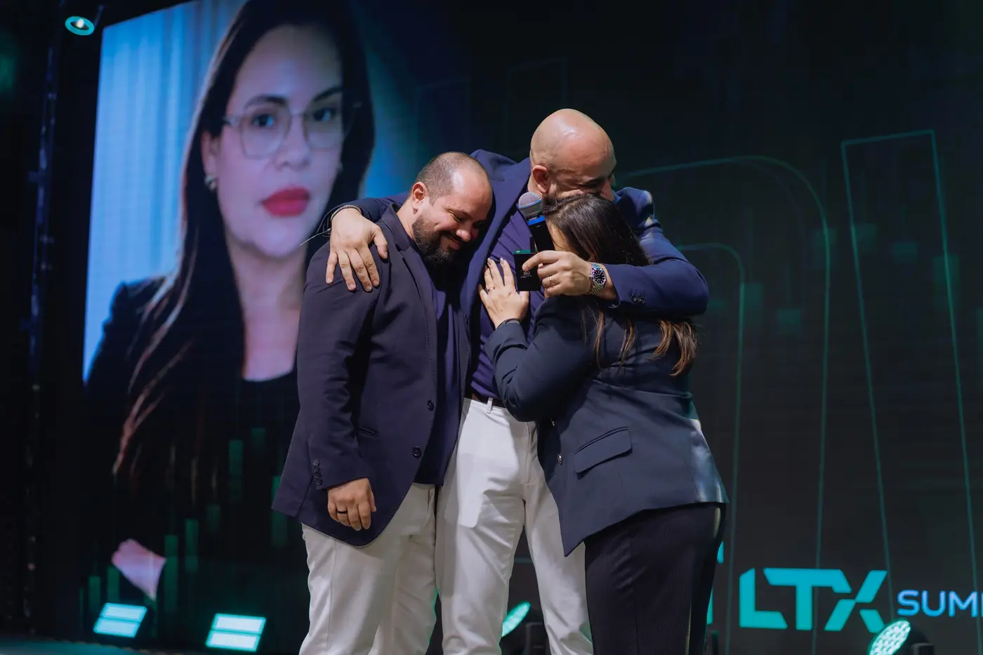 Abraço coletivo no palco com o rosto de uma participante no telão